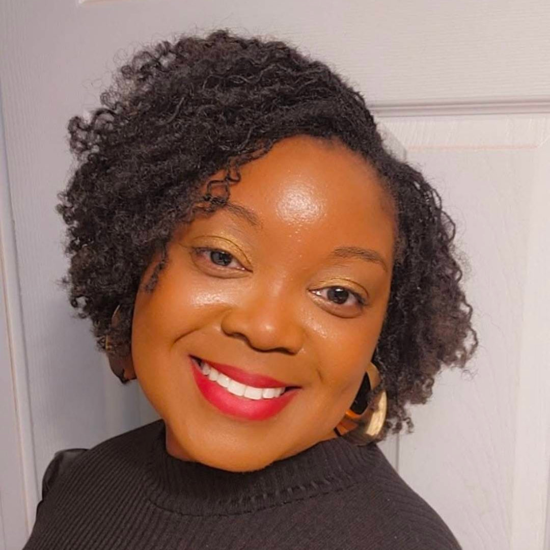 A smiling woman with short, curly black hair wearing a dark ribbed turtleneck sweater and gold hoop earrings, photographed against a light-colored indoor door background.